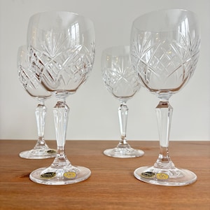 May include: Four clear crystal wine glasses with intricate cut patterns. The glasses have a classic design with a rounded bowl, a faceted stem, and a circular base. The glasses are displayed on a wooden surface.