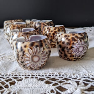 May include: A set of small, round decorative bowls with a leopard print design. Each bowl has a floral pattern in white and pink. The bowls are displayed on a white lace tablecloth.