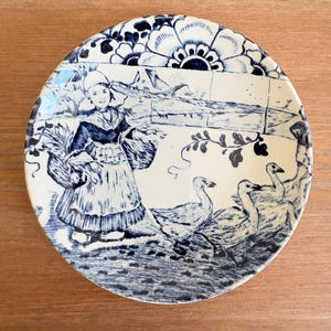 May include: A round, ceramic plate with a blue and white pastoral scene. The design features a woman in traditional attire, geese, and floral patterns. The plate has a diameter of approximately 8 inches.