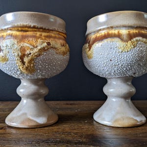 May include: Two handmade ceramic goblets. The goblets have a bulbous body with a textured, speckled surface in shades of white and gray. The rim features a band of brown and gold. The stem is a light beige color.