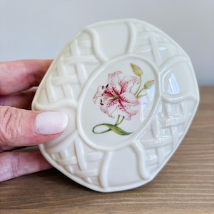 Könnte beinhalten: Eine cremefarbene, ovale Keramikschale mit einer erhabenen Korbgeflecht-Struktur am Rand und einer zentralen Abbildung einer rosa Lilie mit grünen Stielen und Knospen. Die Oberfläche weist ein glänzendes Finish und abgerundete Kanten auf.