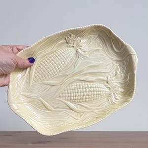 Puede incluir: Plato de cerámica color crema con diseño en relieve de maíz y hojas. El plato rectangular presenta dos mazorcas de maíz y detalles de hojas fluidas, ideal para servir alimentos.