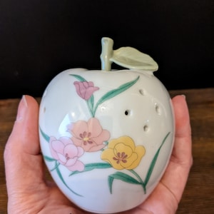 Puede incluir: Un objeto decorativo con forma de manzana de cerámica blanca con diseños florales en rosa, melocotón y amarillo. La manzana tiene un tallo y una hoja verdes, y varios agujeros pequeños. El artículo se sostiene en una mano.