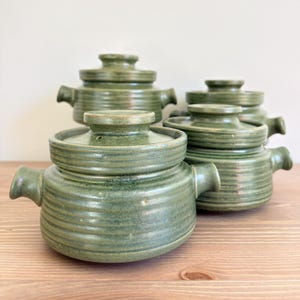 Può includere: Un set di tre pentole in ceramica verde con coperchi abbinati, ciascuna caratterizzata da texture a coste orizzontali e due manici laterali. Le pentole presentano una finitura lucida con sottili variazioni nello smalto verde e forma rotonda.