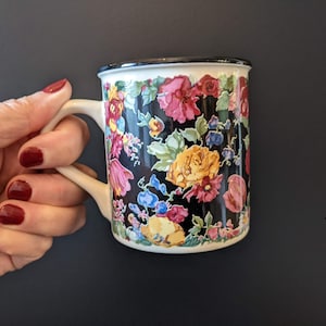 Può includere: Una tazza a fantasia floreale su sfondo nero, con fiori colorati nei toni del rosa, giallo e blu. La tazza ha un bordo e un manico bianchi. La tazza è tenuta in mano con lo smalto rosso.
