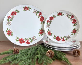 May include: A set of white ceramic plates featuring a circular pattern of red poinsettia flowers, green holly leaves, and small red berries. The plates have a thin gold rim and are arranged with one large plate and a stack of three smaller plates.