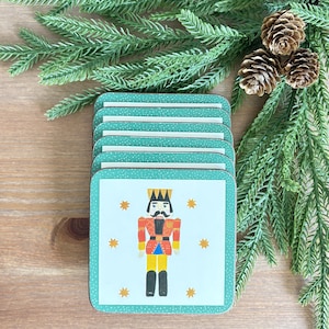 May include: A stack of square coasters featuring a white centre with a graphic of a nutcracker in a red, yellow, and black uniform, surrounded by small yellow stars. The border is teal with a white speckled pattern on the edges.