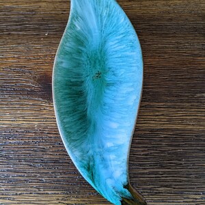 Puede incluir: Un plato decorativo largo con forma de hoja y acabado brillante. El plato presenta una mezcla de tonos turquesa y verde, con un acento marrón oscuro en la base. El diseño tiene un efecto marmolado, agregando interés visual a la pieza de cerámica.