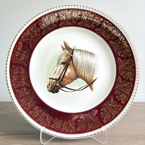 May include: A round ceramic decorative plate featuring a central illustration of a brown horse head wearing a bridle. The plate has a wide, deep red border decorated with intricate gold filigree patterns and a scalloped, gold-trimmed edge.