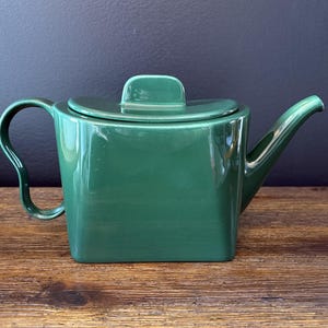 Può includere: Una teiera quadrata in ceramica verde smeraldo con coperchio abbinato e manico curvo. La teiera ha un beccuccio e una piccola maniglia sul coperchio. La teiera è appoggiata su una superficie di legno.
