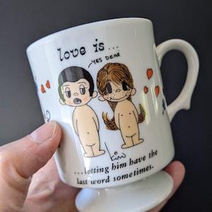 May include: White ceramic mug with a handle and a pedestal base. The mug features a cartoon couple illustration and the text "love is... YES DEAR ... letting him have the last word sometimes."