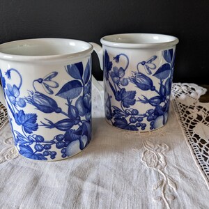 May include: Two white ceramic mugs with blue floral patterns. The mugs have a cylindrical shape with a handle and a white rim. The floral design includes leaves, berries, and flowers. The mugs are placed on a white lace doily.