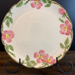 Puede incluir: Un plato de cerámica color crema con borde festoneado, adornado con diseños florales rosas y hojas verdes. El plato está sostenido por un soporte de metal negro, mostrando una estética decorativa y elegante. El patrón floral incluye flores rosas.
