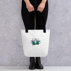 May include: White tote bag with black handles featuring a green and silver plant design inside a black square. The bag is held by a person wearing black pants and boots.