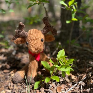 May include: A handmade brown knitted reindeer with dark brown antlers and a red bow tie. The reindeer is sitting on a bed of leaves and twigs, with green foliage in the background. The toy is a whimsical woodland creature.