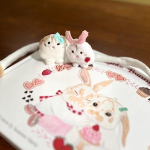 May include: Two small plush bunny figures, one white with pink ears and a red heart, the other beige with a blue bow. They sit on a white tray with a bunny illustration, strawberries, and hearts.