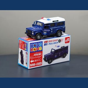 Peut inclure: Un camion de police jouet bleu en métal moulé sous pression avec un toit blanc, posé sur une boîte. La boîte porte les mots "HONG KONG" et "MM-095", ainsi qu'une illustration du camion. Le camion porte le mot "POLICE" sur le toit.