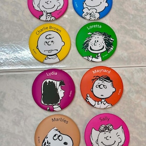 May include: A set of eight circular pinback buttons featuring Peanuts characters. The buttons are colored pink, blue, yellow, green, purple, orange, and tan. Each button displays a character illustration and a name: Sally, Linus, Charlie Brown, Loretta, Lydia, Maynard, and Marbles. Each button includes the text PNTS.