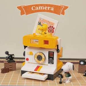 May include: A yellow and white toy camera with a photo of a yellow bird and a paw print. The camera has a red, yellow, and orange stripe. A Snoopy figurine stands next to the camera. The word "Camera" is on a banner above the camera.