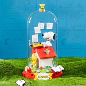 May include: A Snoopy-themed decorative piece featuring Snoopy at a typewriter on top of his red doghouse, enclosed in a clear dome. The base is red with the word "PEANUTS" in yellow. Includes a yellow Woodstock figure.