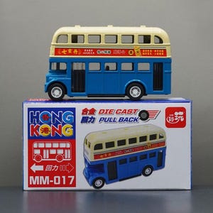 May include: A blue and cream-colored double-decker bus toy with black wheels sits atop a box. The box features the words "HONG KONG" and an illustration of the bus. The toy bus has the words "DIE CAST PULL BACK" on the box.