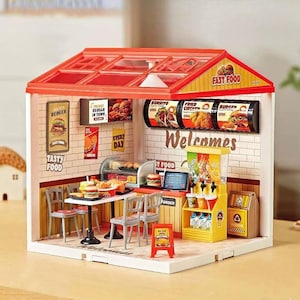 May include: A miniature fast food restaurant model with a red roof and white brick walls. The interior features tables, chairs, and food displays. Signs read "Burger," "Burrito," and "Fried Chicken." The text "Tasty Food" and "Welcomes" are also visible.