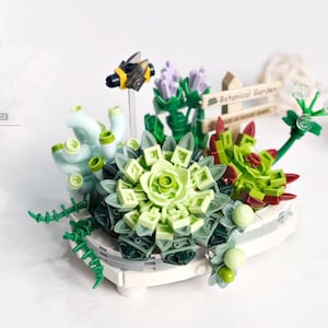 May include: A miniature botanical garden scene constructed from building blocks. The arrangement features various succulents in shades of green, with a small bee and a sign that reads "Botanical Garden". The set includes 389 pieces.