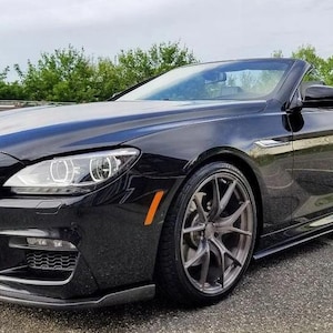 Może przedstawiać: Czarny kabriolet o eleganckim designie zaparkowany na utwardzonej nawierzchni. Samochód ma niski profil, felgi aluminiowe i czarną osłonę chłodnicy. Reflektory są jasne, a samochód wydaje się być w doskonałym stanie.