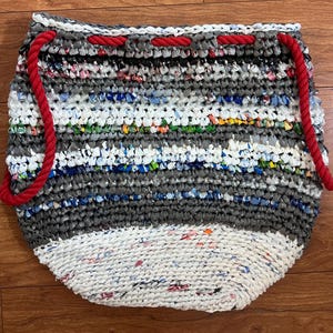 May include: Handmade tote bag crafted from recycled plastic, featuring a striped pattern in shades of gray, white, and colorful accents. The bag has red rope handles and a drawstring closure. A sustainable and eco-friendly accessory.