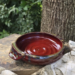 May include: A handmade, round ceramic bowl with a deep red interior and a dark, glazed exterior. The bowl has two small handles and a spiral pattern in the center. The rim has a green and blue glaze. A rustic, decorative piece.