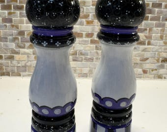May include: Pair of decorative salt and pepper grinders. The grinders have a black top with white floral accents, a purple band, and a white body with black and purple details. The base has vertical stripes.