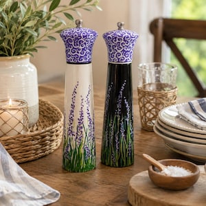 May include: Two tall, tapered salt and pepper mills featuring hand-painted lavender floral designs. One mill has a white base, and the other has a black base. Both mills are topped with purple, swirl-patterned caps and silver-toned metal knobs.