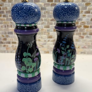 Puede incluir: Par de molinillos decorativos de sal y pimienta. Los molinillos son negros con diseños florales en verde, morado y azul. Las tapas son azules con un patrón de remolino y los molinillos tienen tapas doradas.
