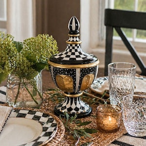 May include: A decorative black and white checkered jar with a gold base and accents, topped with a black and white finial. The jar sits on a table with a woven placemat, checkered plates, and clear drinking glasses. A lit candle in a small glass holder adds warmth.