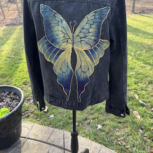Può includere: Giacca di jeans scura con un grande disegno di farfalla sul retro. La farfalla ha ali blu e oro. La giacca è esposta su un manichino nero.