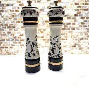 May include: A pair of decorative salt and pepper grinders. The grinders are white with black and gold accents. The tops have a black and white floral design. The base is black with a gold band. The grinders are approximately 10 inches tall.