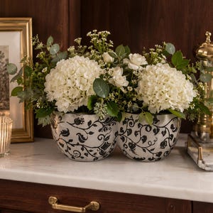 May include: A double-bowl ceramic planter featuring a white base with a black intricate floral and vine pattern. The planter is filled with a lush arrangement of white hydrangea blooms, small white roses, and green eucalyptus foliage.