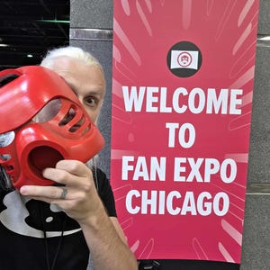 Puede incluir: Una m&aacute;scara de pl&aacute;stico rojo brillante con un dise&ntilde;o angular estilizado que presenta m&uacute;ltiples ranuras de ventilaci&oacute;n y una abertura circular, junto a una pancarta roja vertical con el texto blanco Welcome to Fan Expo Chicago.