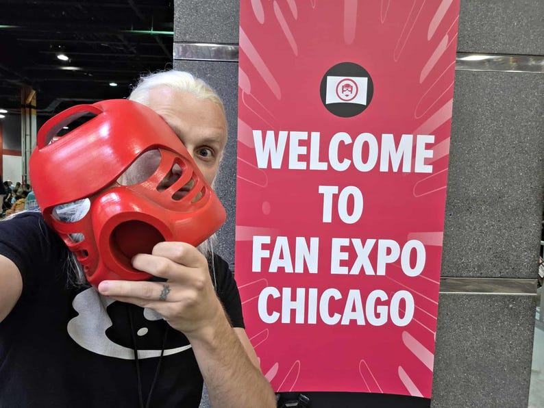 Puede incluir: Una m&aacute;scara de pl&aacute;stico rojo brillante con un dise&ntilde;o angular estilizado que presenta m&uacute;ltiples ranuras de ventilaci&oacute;n y una abertura circular, junto a una pancarta roja vertical con el texto blanco Welcome to Fan Expo Chicago.