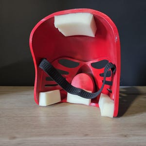 Puede incluir: El interior de un casco rojo con bloques de espuma blanca acolchada adheridos a la carcasa interna. Una correa el&aacute;stica negra ajustable est&aacute; asegurada en el centro, y la estructura incluye m&uacute;ltiples aberturas para los ojos y la nariz.