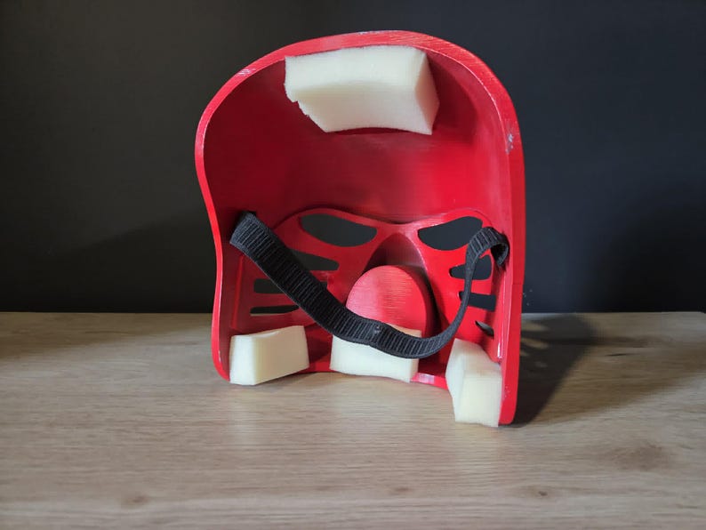 Puede incluir: El interior de un casco rojo con bloques de espuma blanca acolchada adheridos a la carcasa interna. Una correa el&aacute;stica negra ajustable est&aacute; asegurada en el centro, y la estructura incluye m&uacute;ltiples aberturas para los ojos y la nariz.