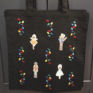 May include: Black tote bag with embroidered colorful Christmas lights and character patches. The bag features a ballerina, a soldier, a nutcracker, and a girl in a white dress. The bag has two black handles.