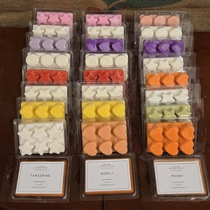 Puede incluir: Filas de ceras perfumadas en varios colores y formas, incluyendo estrellas, corazones y círculos. Cada paquete está etiquetado con un nombre de aroma, como "Tangerine", "Neroli" y "Peony". Las ceras están en envases de plástico transparente.