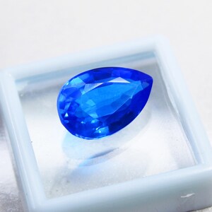 May include: A faceted, pear-shaped, brilliant blue gemstone. The gem is displayed inside a square, white plastic case. The gem's facets reflect light, creating a sparkling effect. The background is a soft, white surface.