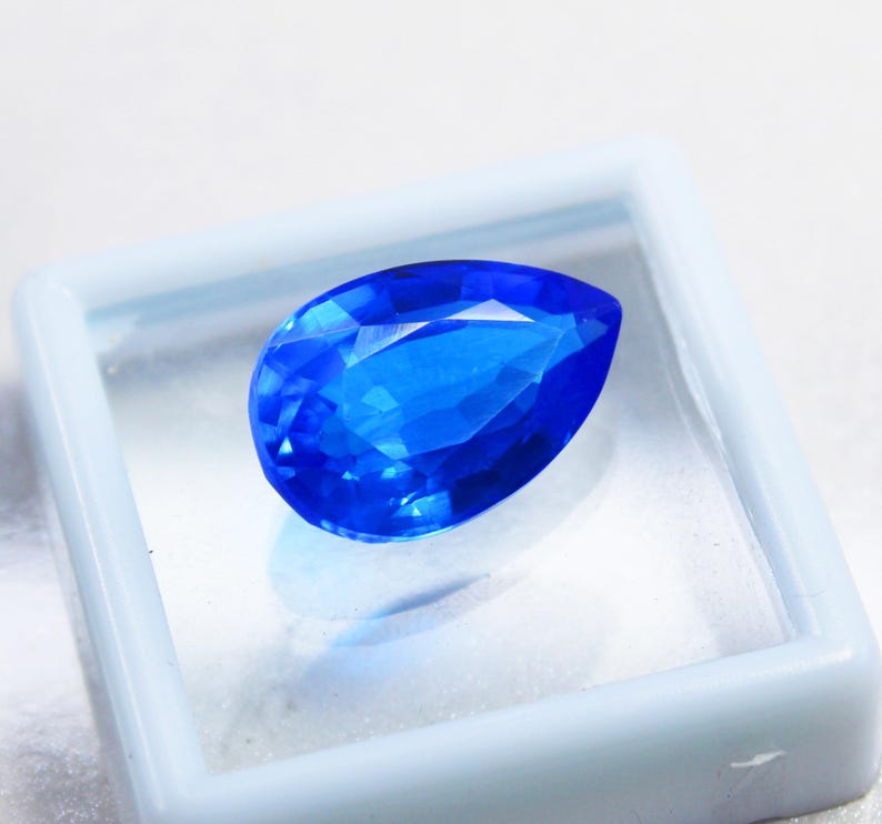 May include: A faceted, pear-shaped, brilliant blue gemstone. The gem is displayed inside a square, white plastic case. The gem's facets reflect light, creating a sparkling effect. The background is a soft, white surface.