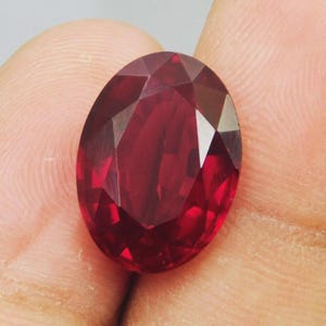 Peut inclure: Une pierre précieuse de taille ovale d'une couleur rouge profonde et saturée avec une surface à facettes qui reflète la lumière. La pierre présente un agencement symétrique de facettes triangulaires et en forme de cerf-volant.