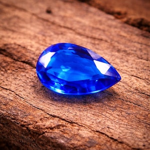 May include: A brilliant, pear-shaped, deep blue gemstone rests on a weathered, brown wooden surface. The gem's facets reflect light, creating bright highlights. The stone's color is a rich, saturated blue, contrasting with the wood's texture.