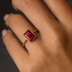Könnte beinhalten: Ein Goldring mit einem rechteckigen, im Smaragdschliff geschliffenen, tiefroten Edelstein in einer glatten Zargenfassung. Die Ringschiene ist dünn und poliert und unterstreicht durch ihr schlichtes Design die leuchtende Farbe und Klarheit.