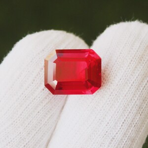 Può includere: Una gemma rettangolare dal taglio a smeraldo con un colore rosso profondo e saturo. La pietra presenta bordi sfaccettati precisi che riflettono la luce, mostrando una qualità chiara e trasparente e una consistenza superficiale lucida e liscia.