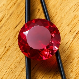 Könnte beinhalten: Ein facettierter, runder Rubin, gehalten von einer schwarzen Pinzette, vor einem hellbraunen Holzuntergrund. Der Rubin hat eine tiefe rote Farbe und reflektiert das Licht von seinen vielen Facetten.